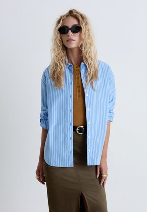 Femme blonde aux cheveux ondulés porte des lunettes de soleil noires, une chemise rayée bleue sur un haut marron boutonné, et une jupe olive fendue à l’avant.