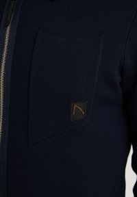 Närbild av en marinblå texturerad jacka med en stor bröstficka och en liten svart lapp med en orange broderad design.