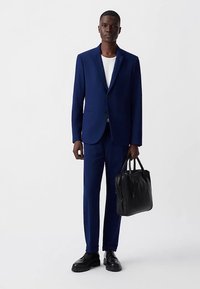 Navyblauw pak met een slanke pasvorm, gestructureerde stof, enkele knoopsluiting, gecombineerd met een wit T-shirt en een zwarte leren tas. Zwarte schoenen.