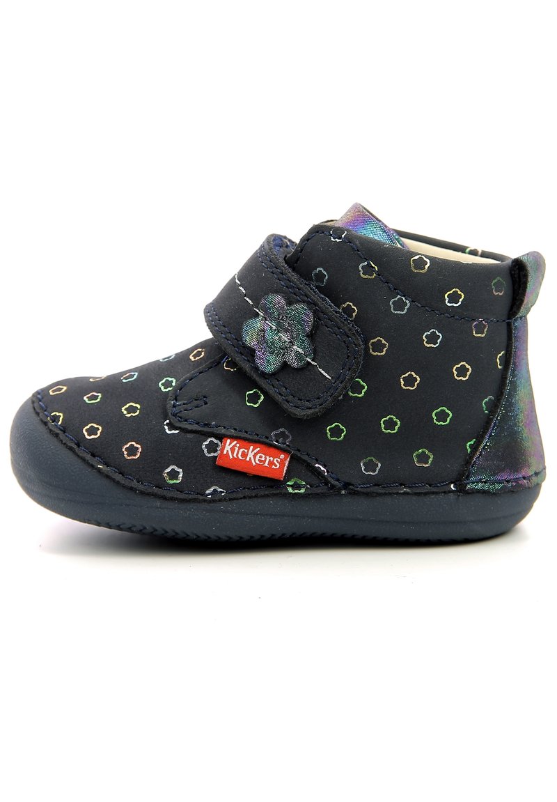 Black high-top boot with multicolored hexagonal pattern, Velcro strap, and a small flower accent. Rubber sole with red Kickers logo.