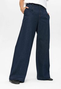 Person wearing dark blue wide-leg denim jeans with hands in pockets and black woven pointed shoes on a white background.