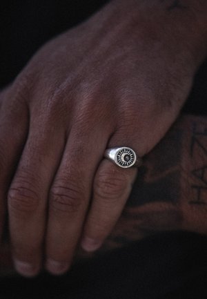 Sculpted silver ring featuring a central black stone surrounded by sunburst detailing. The hand has visible texture and tattoos.