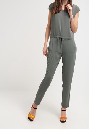 Combinaison vert olive à manches courtes, avec taille à cordon, jambes fuselées, associée à des sandales à talons hauts bicolores orange et noir.