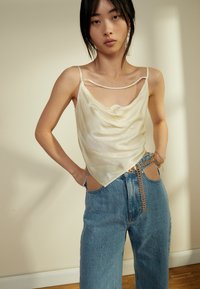 Camisola de cetim cor-de-creme com decote em bico, alças finas, combinada com jeans de denim azul de cintura alta com recortes laterais e detalhe em corrente.