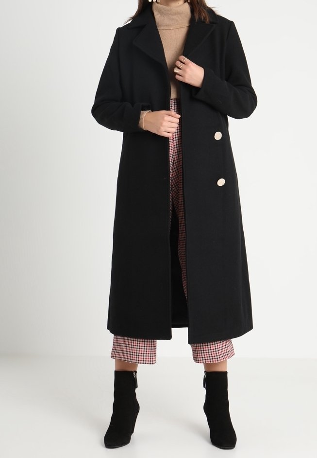 Abrigo de lana negro con un diseño largo, cuello de muesca y dos grandes botones beiges. Lleva puesto encima de un suéter de cuello alto color beige y pantalones a cuadros.