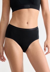 Sezione centrale di donna che indossa biancheria intima nera a vita alta e un reggiseno sportivo nero coordinato, su uno sfondo semplice.