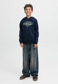Junge lächelt, trägt einen dunkelblauen Kapuzenpulli mit "Jack & Jones"-Aufschrift, weite ausgebleichte Jeans und schwarze Schuhe, steht vor einem schlichten weißen Hintergrund.