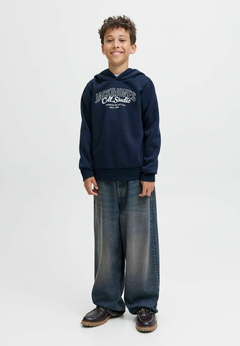 Junge lächelt, trägt einen dunkelblauen Kapuzenpulli mit "Jack & Jones"-Aufschrift, weite ausgebleichte Jeans und schwarze Schuhe, steht vor einem schlichten weißen Hintergrund.