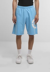 Light blue sweat shorts with an elastic waistband, featuring a large white graphic print on the left leg. Paired with white sneakers and socks.