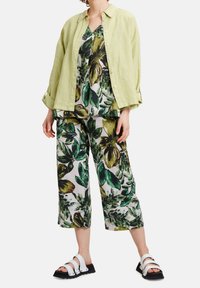 Zelená lněná košile na knoflíky přes květinový top a volné zkrácené kalhoty v odstínech zelené a žluté, doplněné bílými sandály.