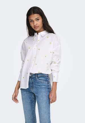 Naine kannab valget nööbitavat särki väikeste kollaste lillemustriga ja siniseid teksaseid, seisab ühevärvilise heleda tausta ees.