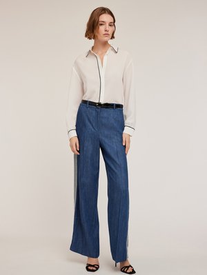 Witte blouse met lange mouwen en zwarte afwerking, gecombineerd met donkerblauwe brede broek met lichtgrijze zijpanelen. Zwarte riem en hakken.