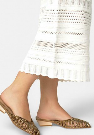 Pies calzando tacones marrones con tachuelas, de punta y sin cierre, combinados con una falda blanca hasta la rodilla con patrones de encaje texturizados.