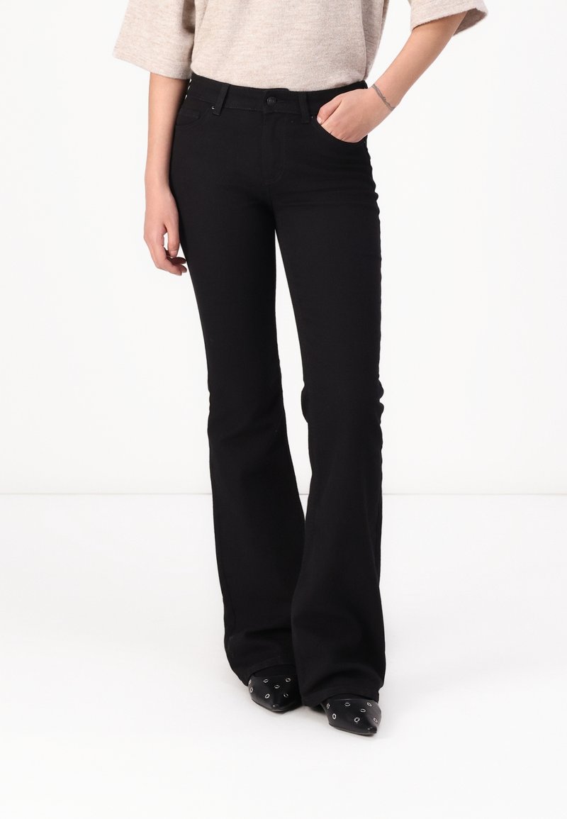 Personne portant un pantalon noir évasé taille haute avec poches avant, associé à des chaussures noires pointues avec œillets métalliques.
