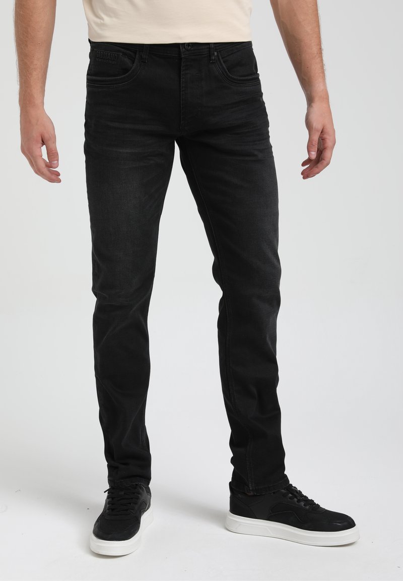 Homme portant un jean noir slim fit et des baskets noires avec des semelles blanches, debout sur un fond uni.