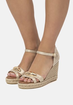 Sandalias de cuña doradas con suela de yute trenzado. La parte superior presenta detalles metálicos y dos correas texturizadas, con una correa en el tobillo.