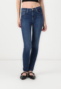 Skinny blue jeans with a mid-rise waist, five-pocket design, and subtle stitching details. Worn with black flats featuring metal accents.