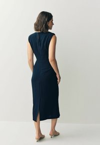 Marinblå, figursydd ärmlös klänning med slits bak och subtil fodring. Slät textur och elegant design framhäver dess strömlinjeformade siluett.