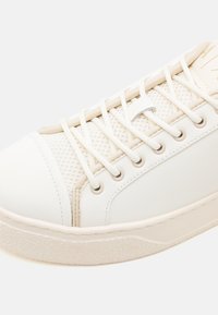 Valge tennis, millel on tekstureeritud võrgupind, siledad nahast detailid, ühte tooni paelad ja kummist tald, millel on veidi paksem kuju.