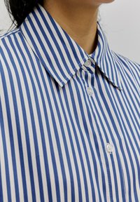 Gestreifte Hemdbluse mit blau-weißen Streifen, klassischem Kragen und weißen Knöpfen an der Vorderseite. Baumwollstoff mit glatter Textur.