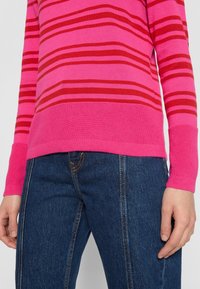 Maglione a righe rosa e rosso in tessuto knit, con polsini e orlo a coste, abbinato a jeans in denim blu scuro con dettagli di cucitura arancione.