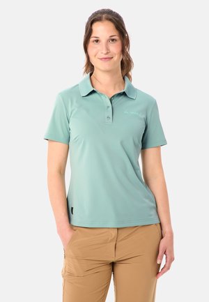 Frau trägt ein hellgrünes, kurzärmeliges Poloshirt und beige Hose, steht mit einer Hand in der Tasche vor einem einfarbigen Hintergrund.