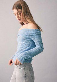 Tânără îmbrăcată cu un pulover tricotat de culoare albastru deschis, cu umerii descoperiți, și blugi gri deschis, privind în jos pe un fundal simplu.