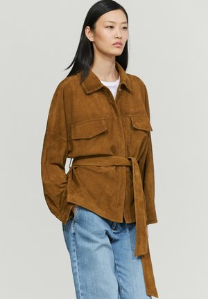 Frau trägt eine braune Wildlederjacke mit Gürtel und Klappentaschen sowie hellblaue Jeans vor hellem grauem Hintergrund.