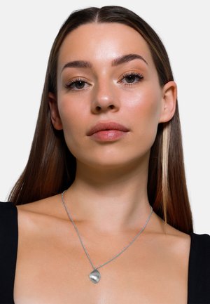 Zilveren ketting met een getextureerde bladhanger op een delicaate schakel, gedragen tegen een zwarte top, die het reflecterende oppervlak laat zien.