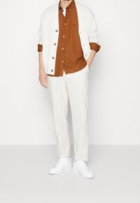 Cardigan bianco con bottoni grandi, camicia marrone con bottoni e pantaloni crema. Abbinati a sneakers bianche. Look semplice e casual con colori neutri.