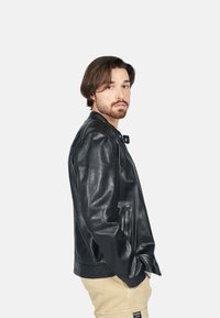 Svart läderjacka med en mjuk textur, dragkedjefickor och en klassisk krage. Modellen bär ljusa khakibyxor och står vänd åt vänster.