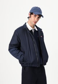 Jachetă albastră închisă, cu fermoar, cu guler înalt, cămașă polo albăbuttonată dedesubt și o șapcă albastră asortată. Are buzunare laterale și manșete elastice.