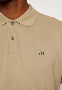 Camisola polo bege feita de tecido texturizado, com gola, dois botões cinzentos e detalhe bordado verde no lado esquerdo.