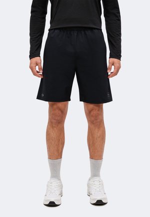 Mand iført sorte sportsshorts, sort langærmet top, grå ankelsokker og hvide sneakers står mod en ensfarvet baggrund.