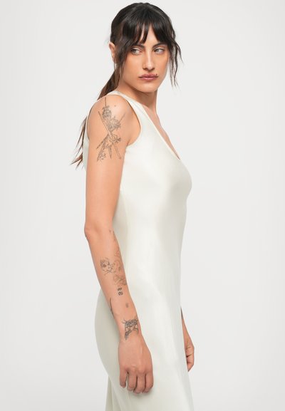 Kvinna med mörkt hår i en vit ärmlös klänning som visar detaljerade tatueringar på sin vänstra arm, tittar åt sin högra sida mot en neutral bakgrund.