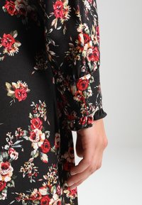 Detalle de un vestido negro floral, con un puño de manga con volantes. La tela presenta vibrantes rosas rojas y blancas con hojas verdes, creando un patrón texturizado.