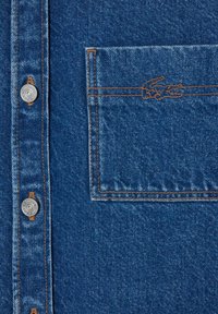 Denim shirt featuring a deep blue color, silver buttons, contrasting orange stitching, and a small embroidered logo on the pocket.