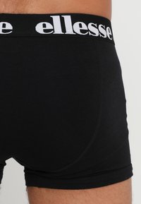 Svarta boxerunderkläder i bomull, med ett brett elastiskt midjeband som har "ellesse"-logotyp i vitt. Slät textur med en åtsittande design.