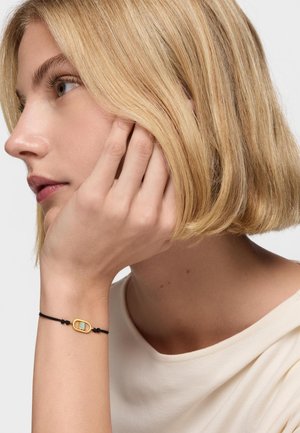 Donna bionda con capelli corti che si tocca il viso, indossando un braccialetto con pietre dorate e verdi su un cordino nero al polso, vestita con una maglietta bianca.