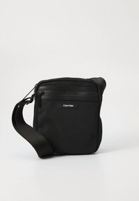 Black Calvin Klein crossbody bag with front zipper pocket and adjustable shoulder strap on a white background.