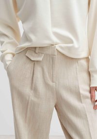 Beige strukturierte Hose mit Falten und flachem Bund, ausgestattet mit einer Seitentasche und einem hellcremefarbenen Langarmoberteil. Einfaches Design und Materialien.