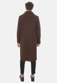 Cappotto di lana marrone con una vestibilità sartoriale, maniche lunghe, colletto a scapola e uno spacco sul retro; abbinato a pantaloni neri slim-fit e scarpe.