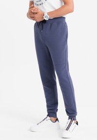 Námořnické modré teplákové kalhoty s ribbovanou texturou, elastickým pasem a nastavitelnou šňůrkou, doplněné bílými teniskami s barevnými pruhy.