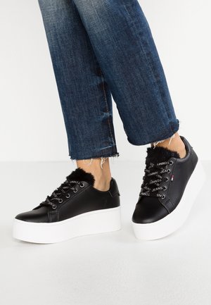 Schwarze Leder-Plateausneaker mit weißer Sohle, ausgestattet mit Kunstfell-Akzenten und markierten Schnürsenkeln. Getragen mit abgenutzten blauen Jeans.