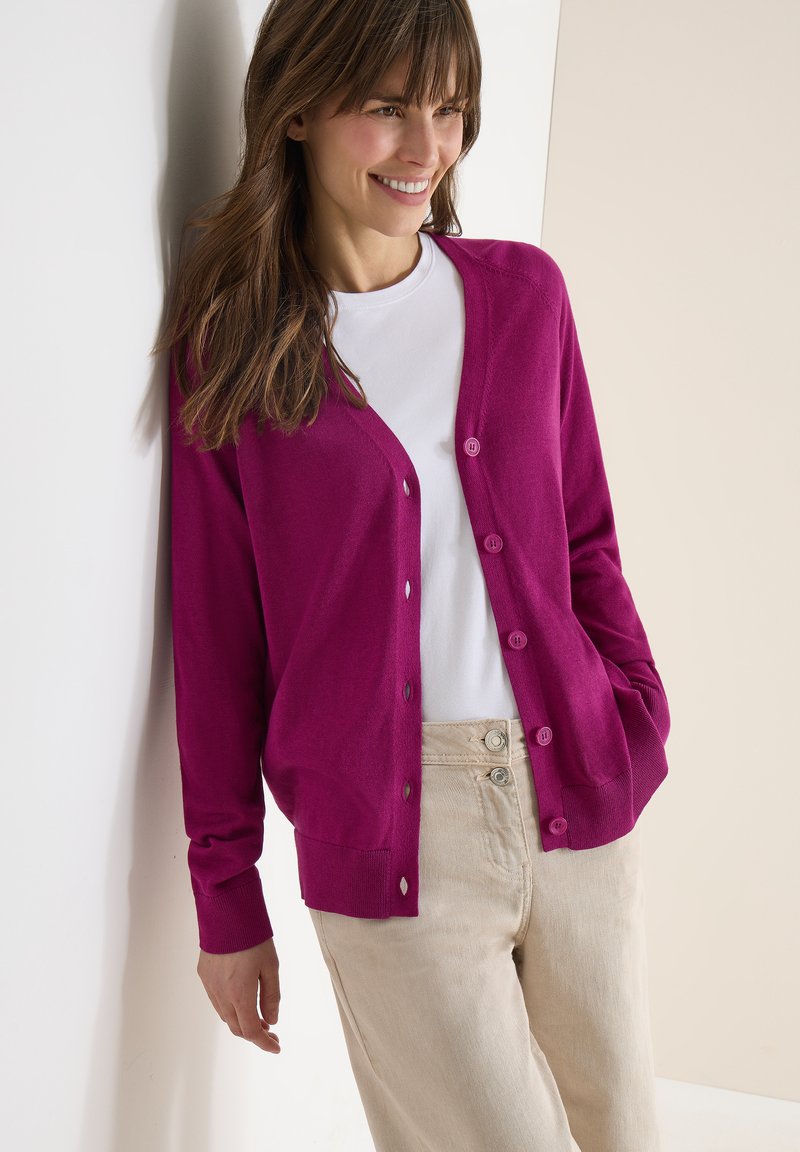 Femme souriante appuyée contre un mur, portant un cardigan violet vif boutonné sur une chemise blanche et un pantalon taille haute beige.