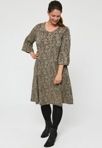 Vrouw die glimlacht, staande in een gebloemde jurk tot de knie met 3/4 mouwen, zwarte panty en zwarte enkellaarsjes op een witte achtergrond.