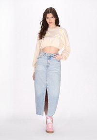 Sweatshirt scurt de culoare crem cu broderie în formă de floare galbenă, asociat cu o fustă maxi din denim de culoare albastră deschisă, cu o fanta frontală, și adidași roz.