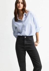 Giovane donna che indossa una camicia a righe blu chiaro con bottoni infilata in jeans neri a vita alta, in piedi con una mano sulla vita su uno sfondo bianco.