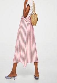 Jupe midi rayée en tissu rouge et blanc avec un détail de taille à nouer. Associée à un haut blanc et un sac en paille tissé. Chaussures grises à bout ouvert.