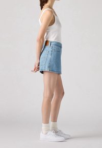 Jupe en denim mini bleu clair, taille haute, avec poche arrière et patch en cuir. Associée à un débardeur blanc et des baskets blanches.